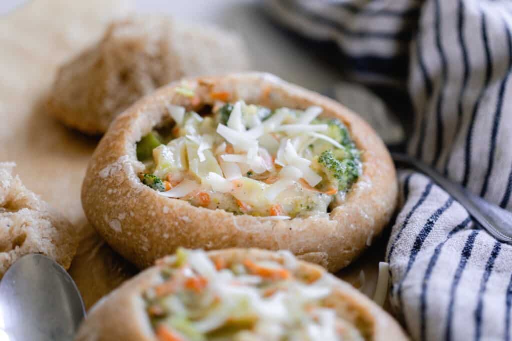 two homemade sourdough bread bowls filled with broccoli cheese soup and topped with more cheese on parchment paper with a tea towel to the right