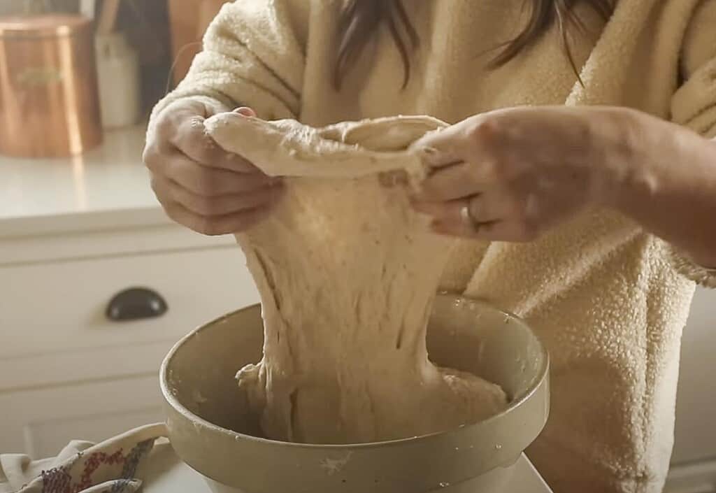 a lady wearing a white sweatshirt stretching dough up above a bowl