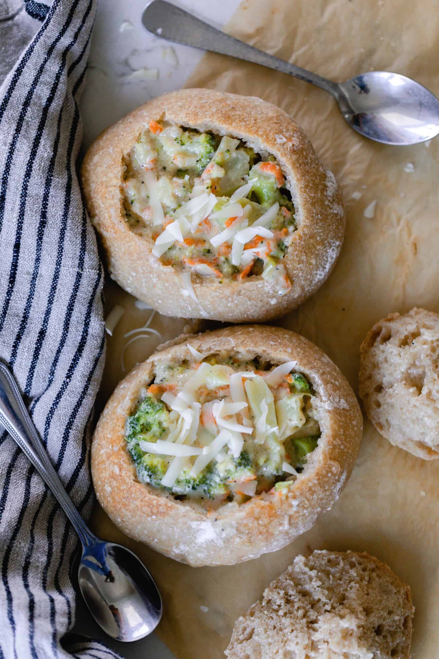 two sourdough bread bowls filled with broccoli cheddar soup and topped with cheese on parchment paper with spoons and a black and white stripped towel around the bread bowls