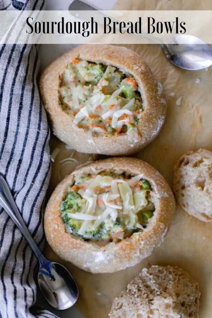 two sourdough bread bowls filled with broccoli cheddar soup and topped with cheese on parchment paper with spoons and a black and white stripped towel around the bread bowls