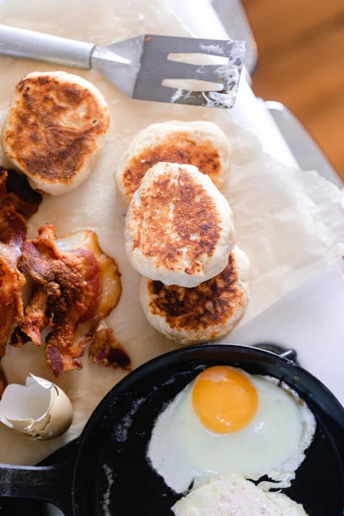 overhead image of sourdough English muffins, crispy bacon, and a cast iron skillet with an sunny side up egg on a wooden table