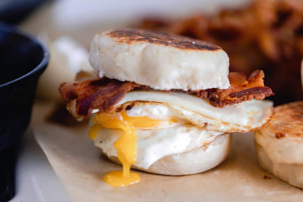 sourdough breakfast sandwich made with sourdough English muffins, thick slices of bacon, cheese, and an over easy egg on parchment paper