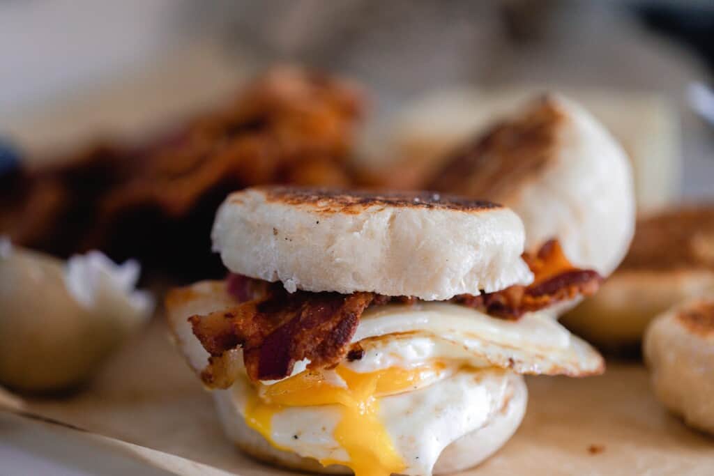 side view of a English muffin breakfast sandwich with bacon, cheese, and egg on parchment paper with more ingredients in the background
