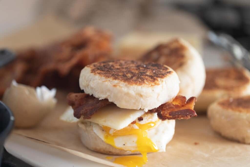 side view of Sourdough English muffin breakfast sandwich with bacon, sharp white cheddar, over easy egg with a broken yolk running down the sandwich. It sits on parchment paper with bacon and English muffins the background