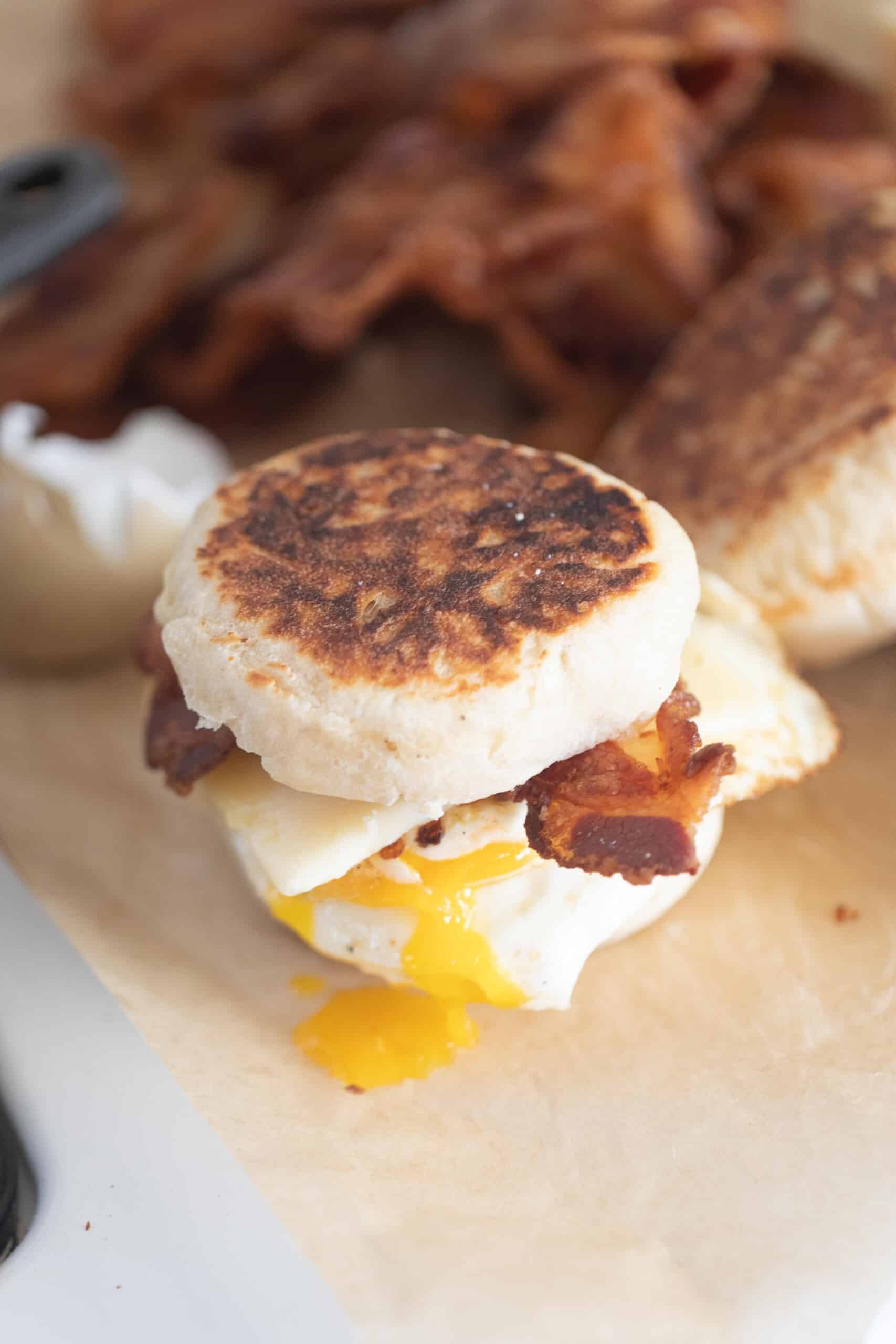 sourdough breakfast sandwich made with English muffins, crispy bacon, sharp cheddar cheese, and an over easy egg with the yolk that has broken and running down the sandwich. The sandwich sits on parchment paper with more ingredients in the background