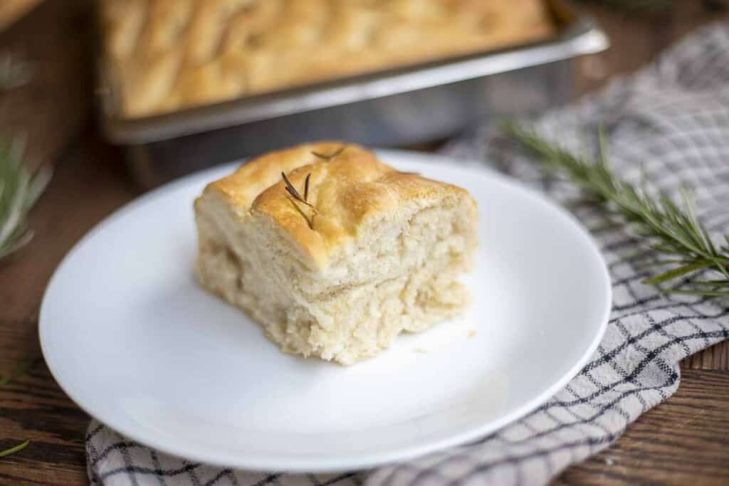 piece of sourdough focaccia made with sourdough discard on a white plate on a black and white towel with a tray of more focaccia in the background