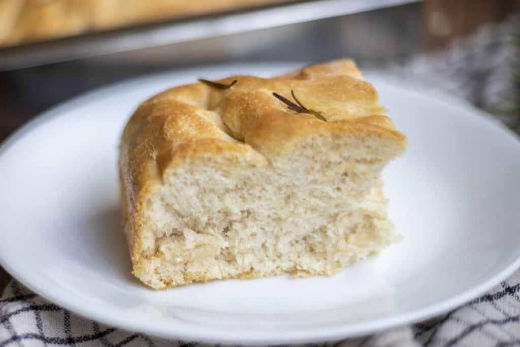 side view of fluffy sourdough discard focaccia bread on a white plate with a tray of more bread in the background