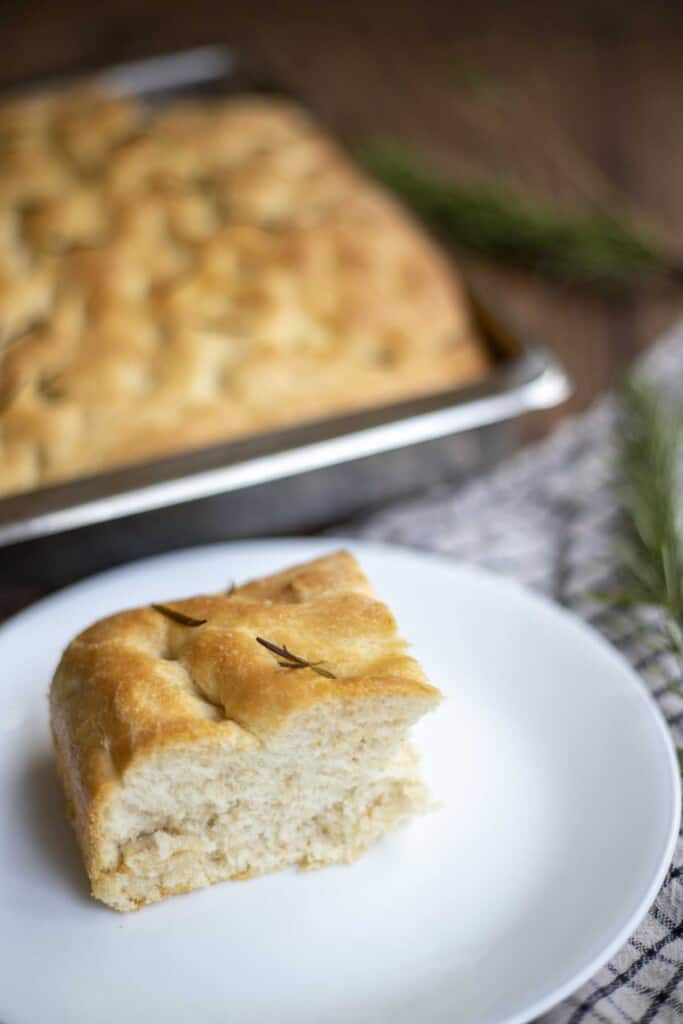 slice of sourdough discard focaccia on a white plate with a tray of more focaccia in the background 