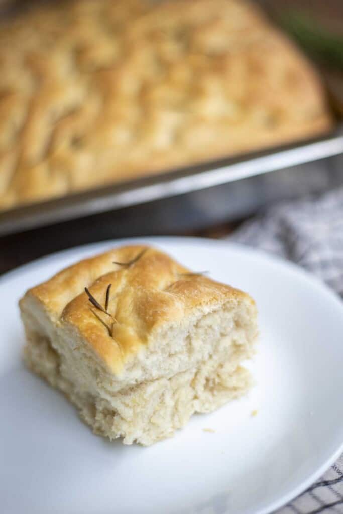 square slice of sourdough discard bread topped with rosemary on a white plate with a baking dish of more focaccia