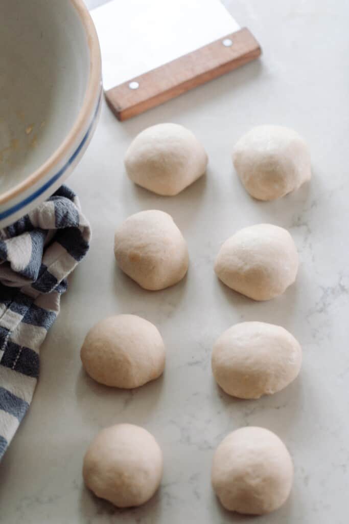 balls of dough on a white quartz countertop with a bench scraper and a bowl in the back left corner