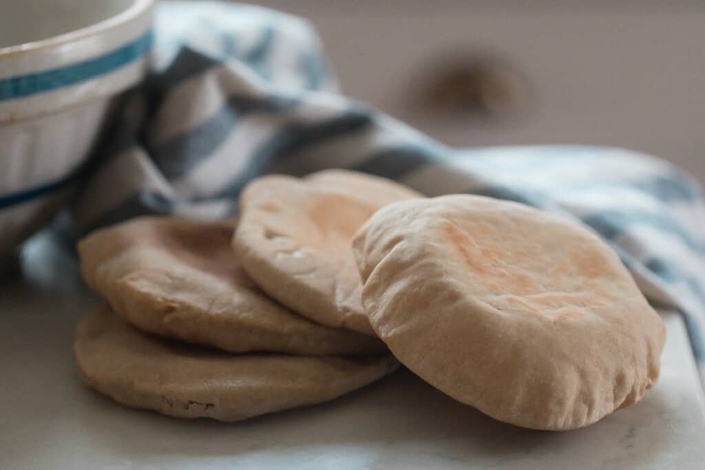 four sourdough pita breads on a white countertop with a white and blue stripped towel and bowl in the background