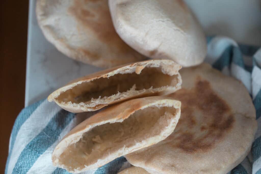 a sourdough pita bread sliced in half revealing a large pocket. More pitas on a white and blue towel surround the opened pita