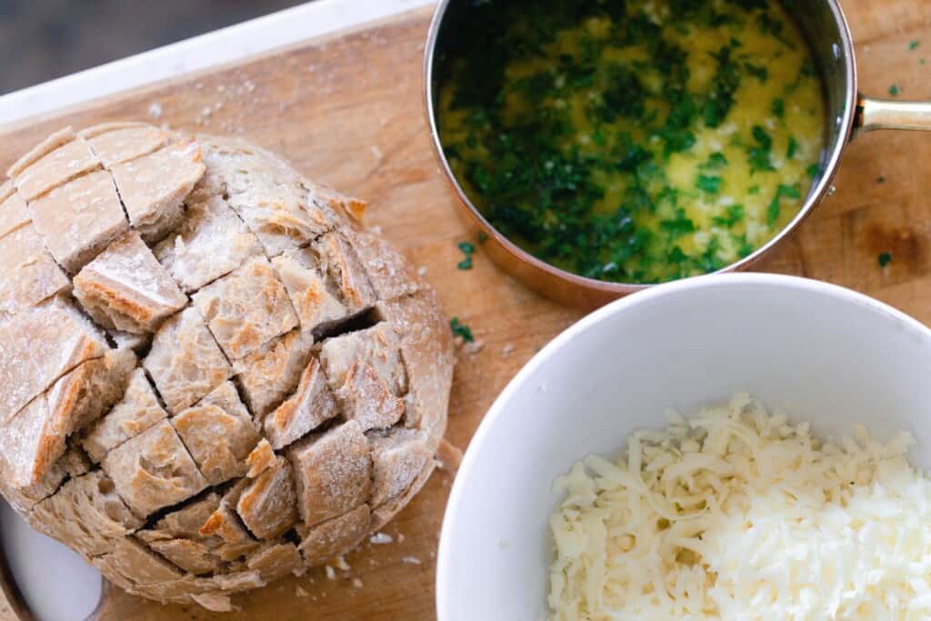 Sourdough boule cut with melted garlic herb butter in bowl and shredded mozzarella in bowl