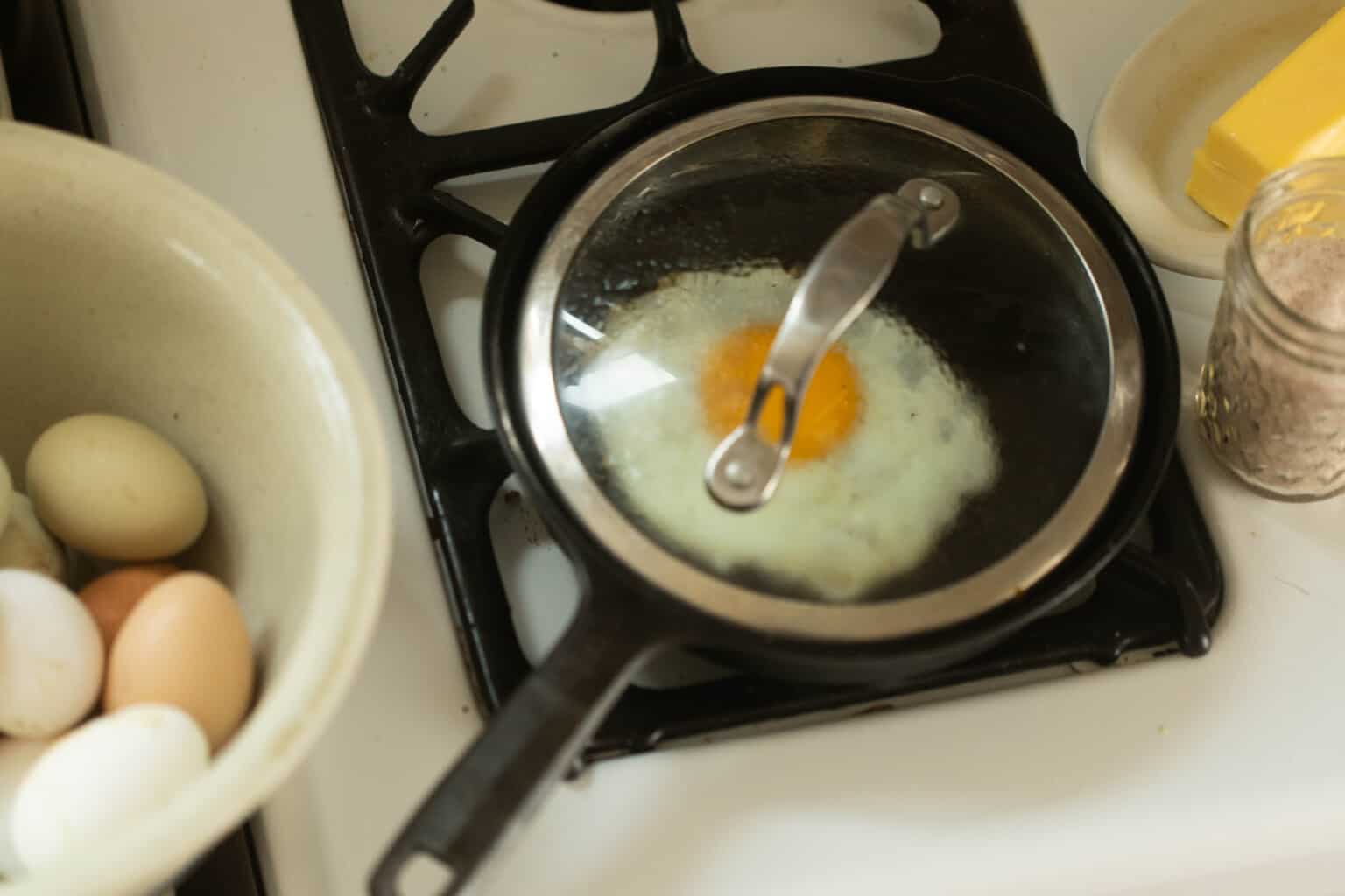 Sunny side up egg cooking in cast iron skillet with glass lid on white stove top.