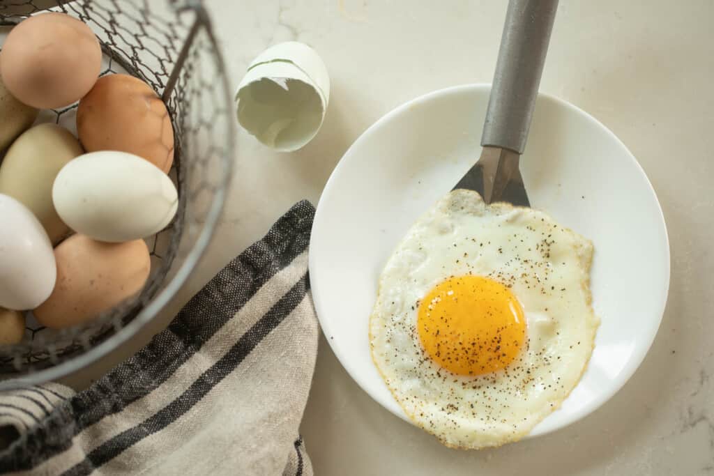 sunny side up egg on spatula being placed on a plate next to a broken egg shell and basket of farm fresh eggs