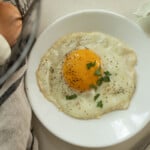 Sunny side up egg on white plate with herbs next to a tea towel.