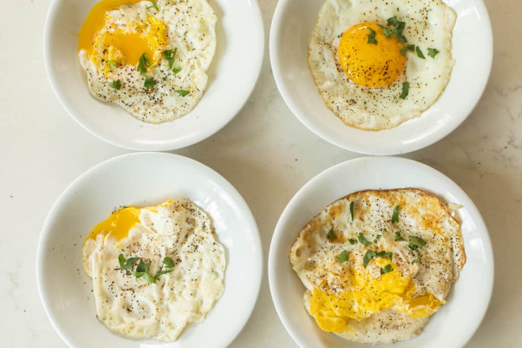 four variations of fried eggs on separate white plates