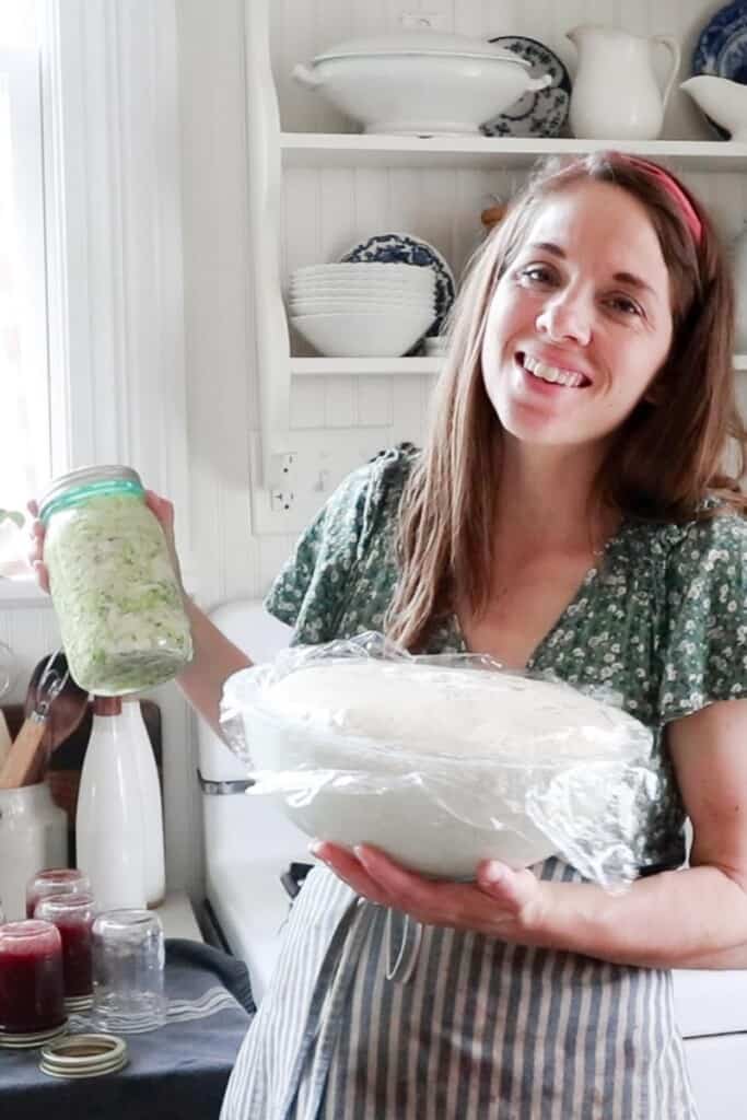 women holding. a blue mason jar of sauerkraut and a bowl of sourdough in her kitchen