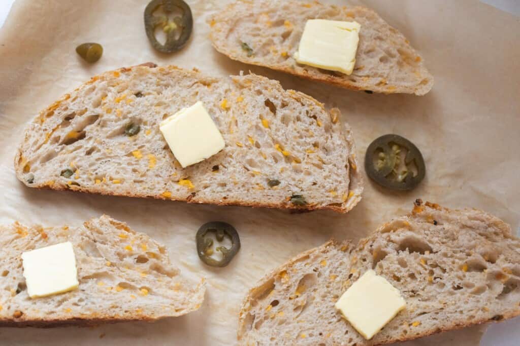 slices of jalapeño cheddar sourdough bread topped with butter on parchment paper with picked jalapeños surrounding the bread slices