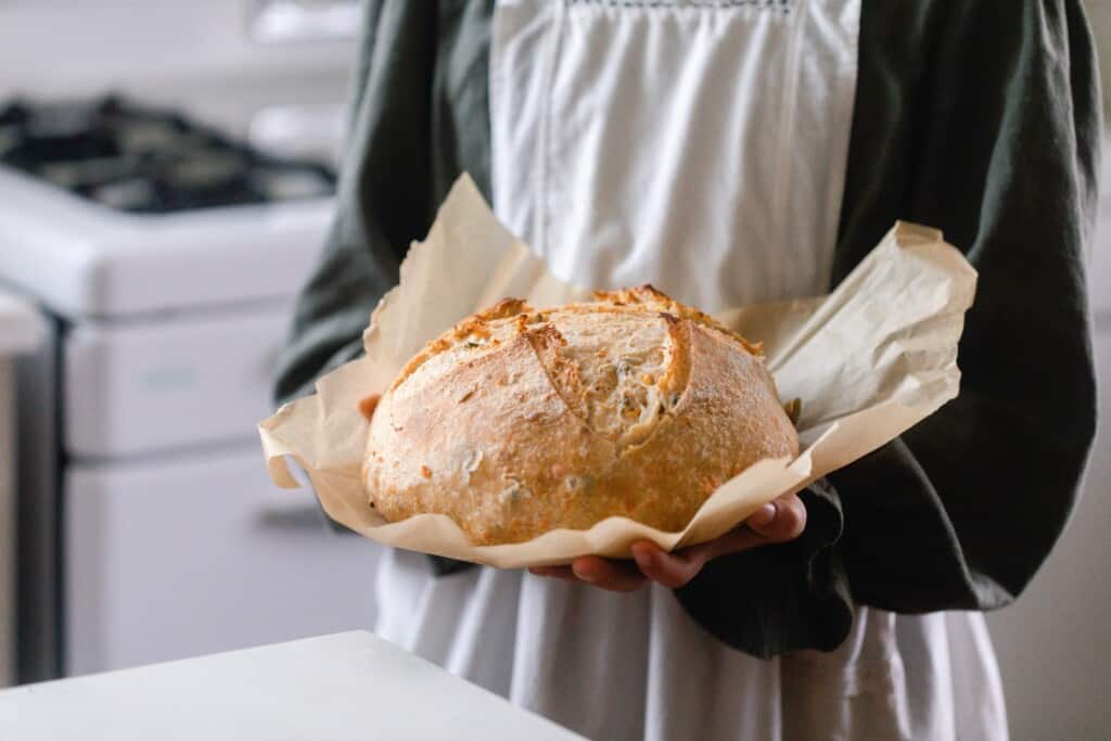 girl in a green dress with a gray apron holding a loaf of cheddar jalapeño sourdough bread on parchment paper