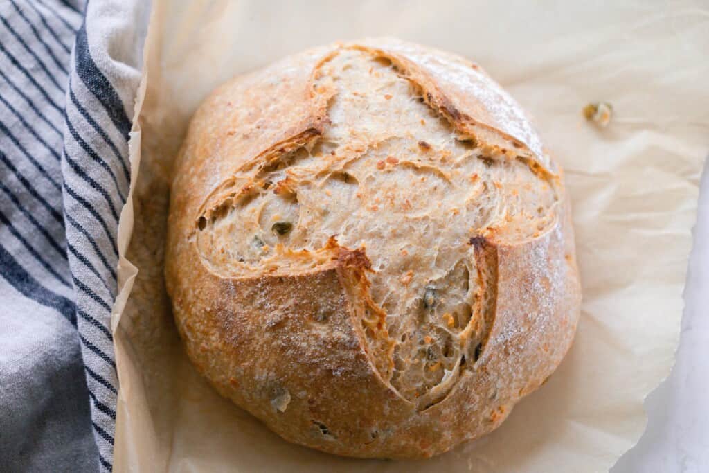 overhead photo of a crusty artisan loaf of cheddar jalapeño sourdough bread on parchment paper with a black and white stripped towel to the left