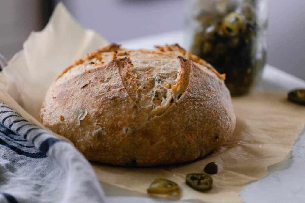 loaf of cheddar jalapeño sourdough bread on parchment paper on a white countertop with pickled jalapeños in the background