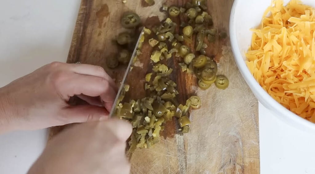 hand chopping pickled jalapeños on a wood cutting board with a bowl of shredded cheese to the left