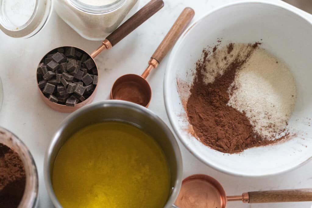 melted butter in a pan with a bowl of coco powder and flour to the right, and copper measuring cups surrounding the bowls