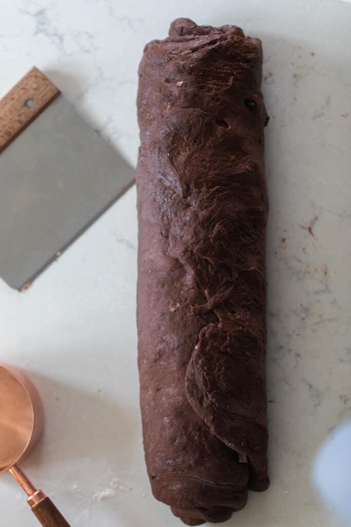 chocolate sourdough sweet roll rolled up on a white countertop with a bench scraper to the left