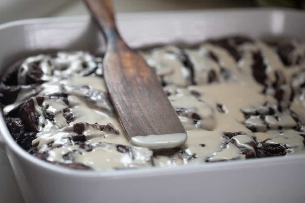 a white baking dish with double chocolate sourdough rolls topped with a vanilla icing with a wooden spatula resting on top