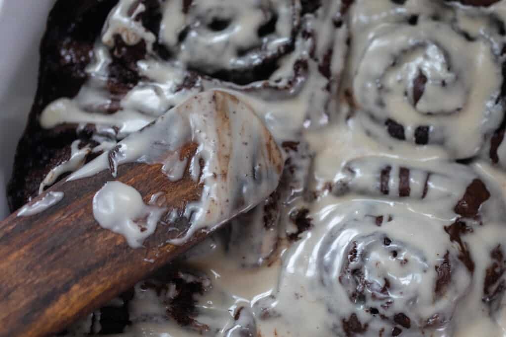 double chocolate sourdough sweet rolls with a vanilla glaze in a baking dish with a wooden spatula