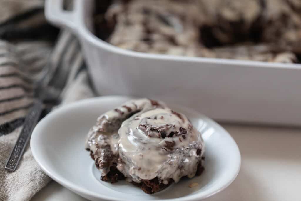 double chocolate sourdough roll with vanilla icing on a white plate with a white baking dish in the background with more rolls