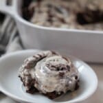 double chocolate sourdough roll with vanilla icing on a white plate with a white baking dish in the background with more rolls