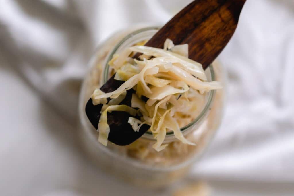 wooden spatula with sauerkraut on top of a jar with more sauerkraut on a white towel