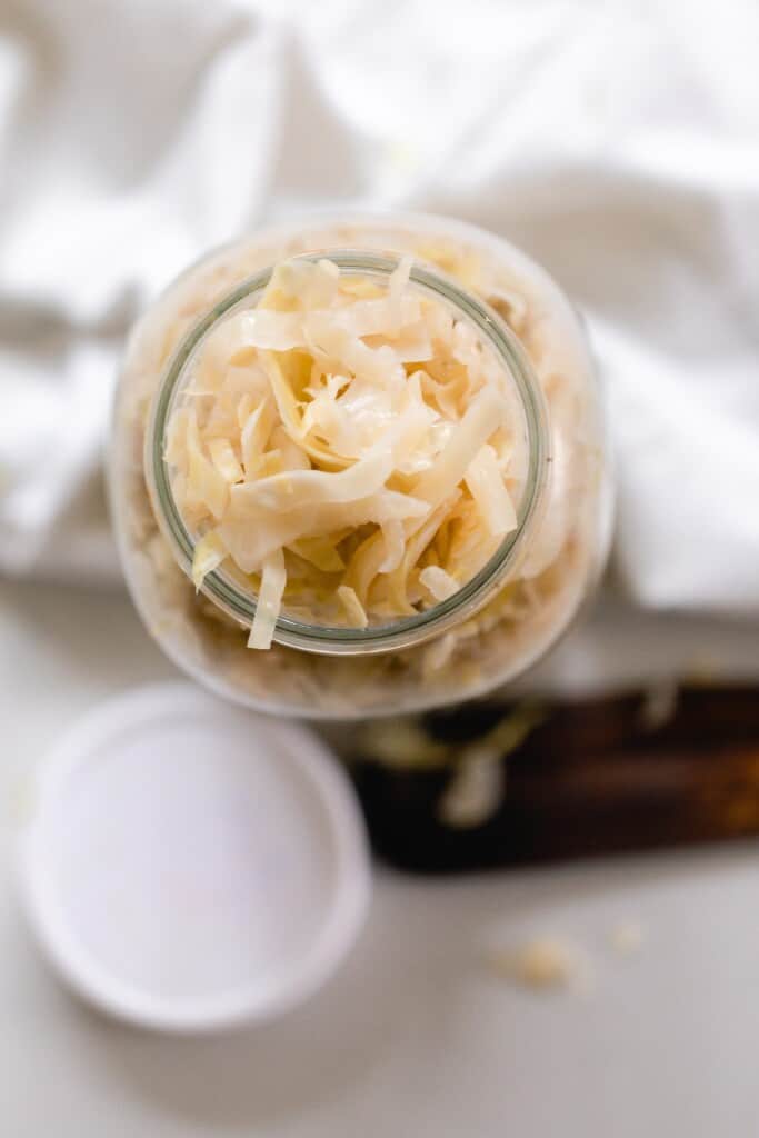 overhead photo of a jar of homemade sauerkraut on a white countertop with a plastic lid and wooden spatula on the counter