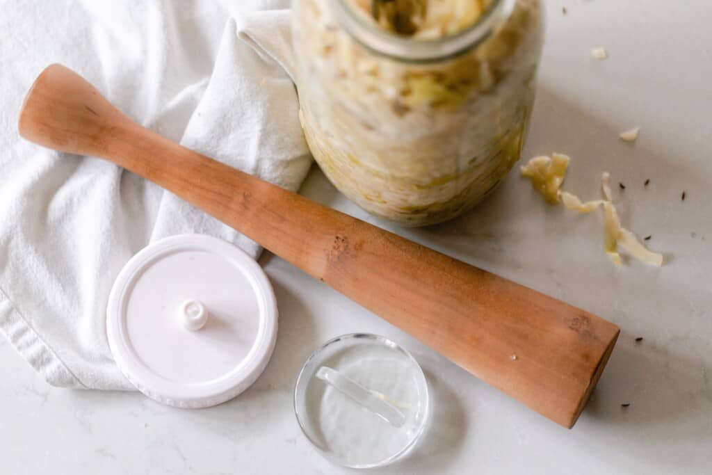 jar of sauerkraut next to a wooden pummel, fermentation lid and glass weight