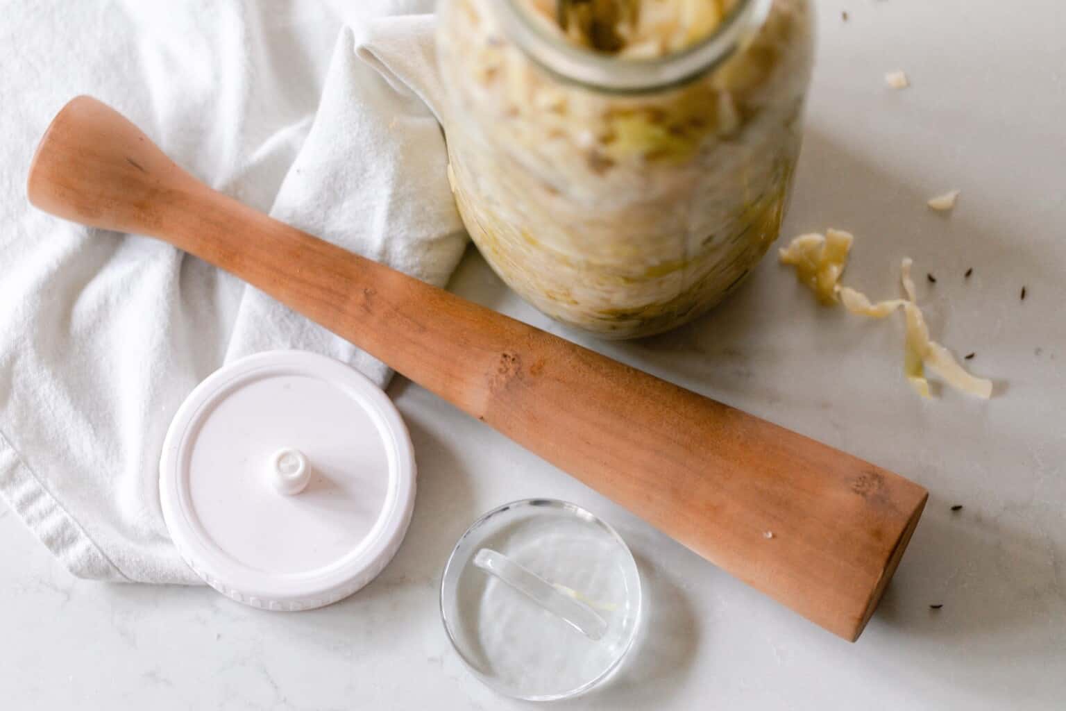 Jar of sauerkraut next to a wooden pummel, fermentation lid, and glass weight.