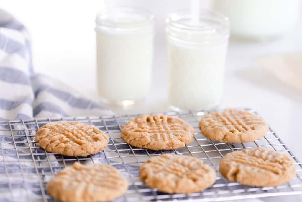 six sourdough peanut butter cookies on a wire rack on top of a blue and white stripped towel with two glasses of milk in the background