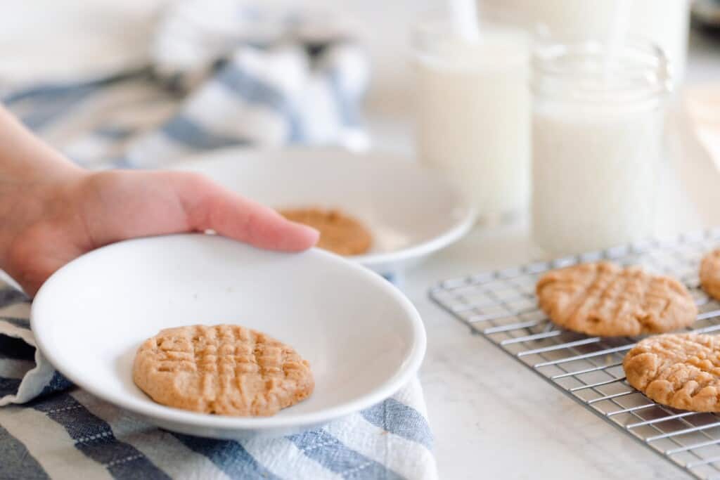 hand holding a plate with a sourdough peanut butter cookie. Glasses of milk and a cooling rack of more cookies are in the background
