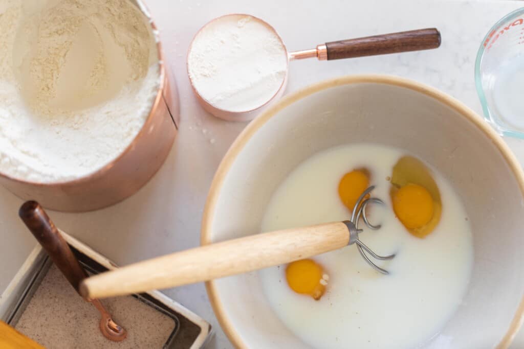 milk and eggs in a bowl with a dough whisk and a cup of flour to the side