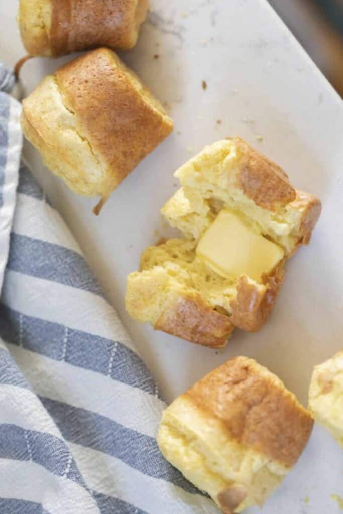 sourdough popover cut in half with a pat of butter on a white countertop surrounded by more popovers and a blue and white stripped towel