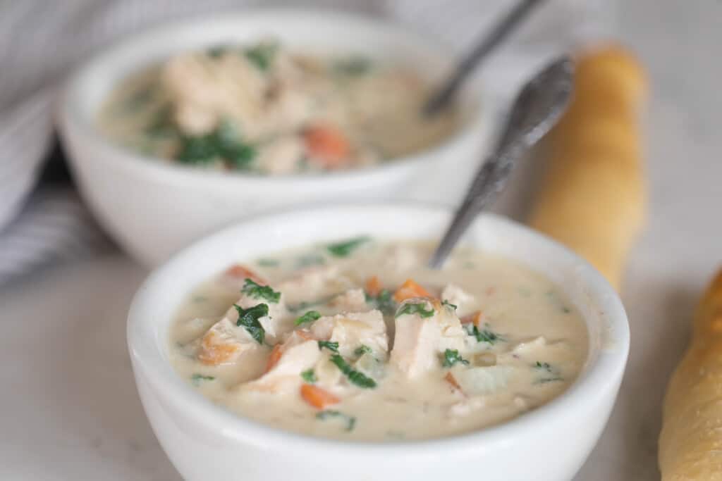 two white bowls filled with chicken kale soup with silver spoons and bread sticks on the side