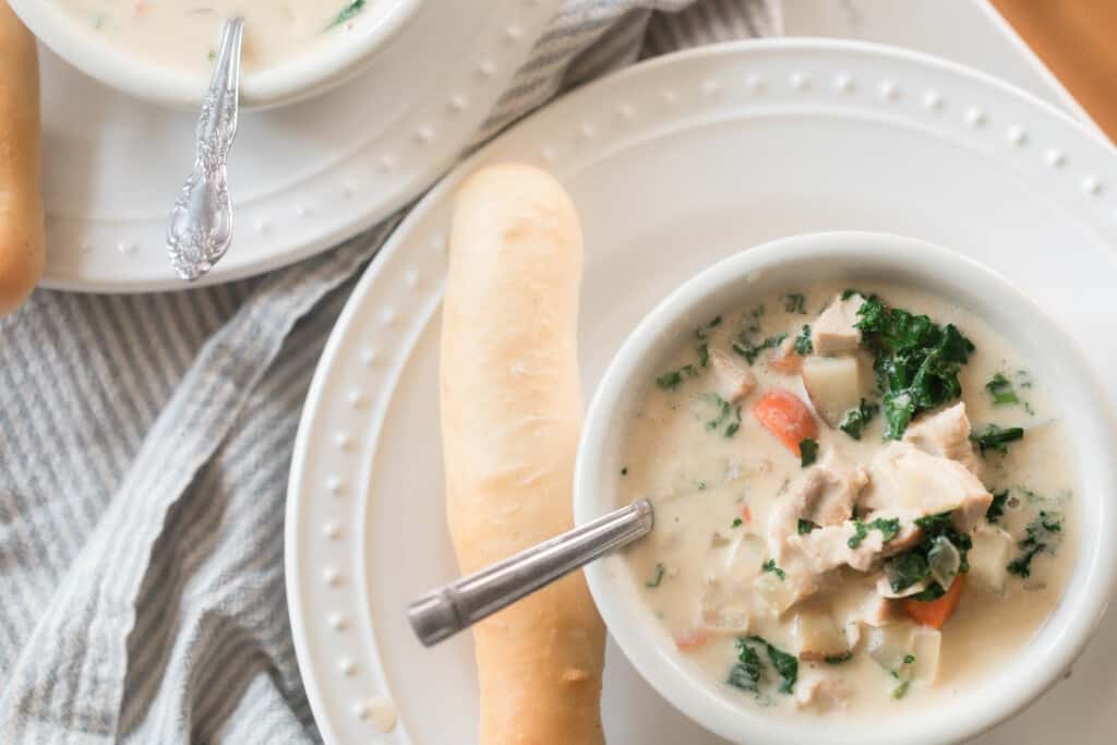 white bowl with chicken kale soup with silver spoon on a white plate with a sourdough breadstick on the side