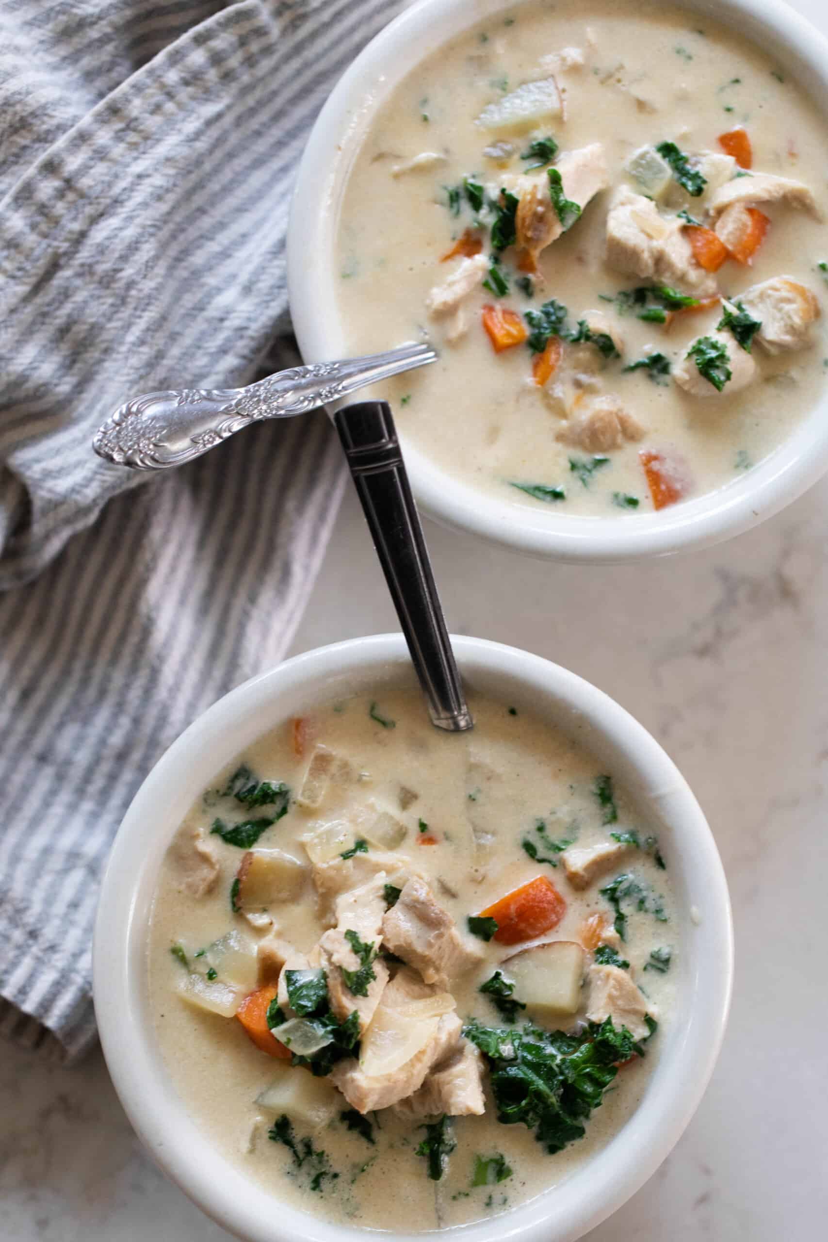 Two bowls of chicken kale soup with a silver spoon in each on a white countertop with a blue and white striped tea towel on the side