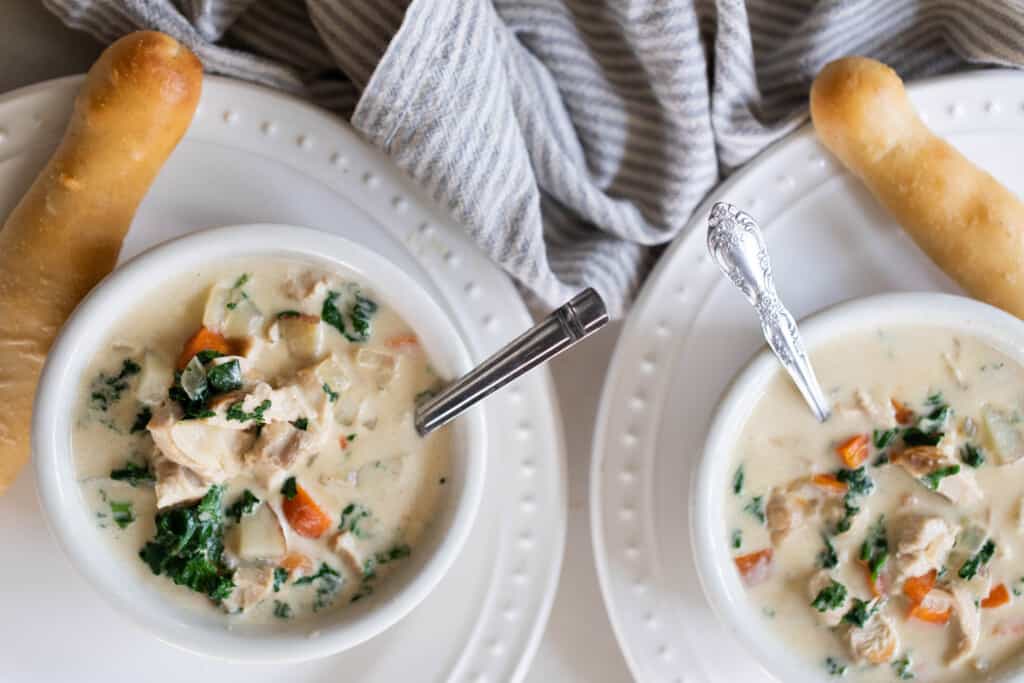 Two bowls of chicken kale soup with spoons on white plates with a sourdough breadstick on each. 