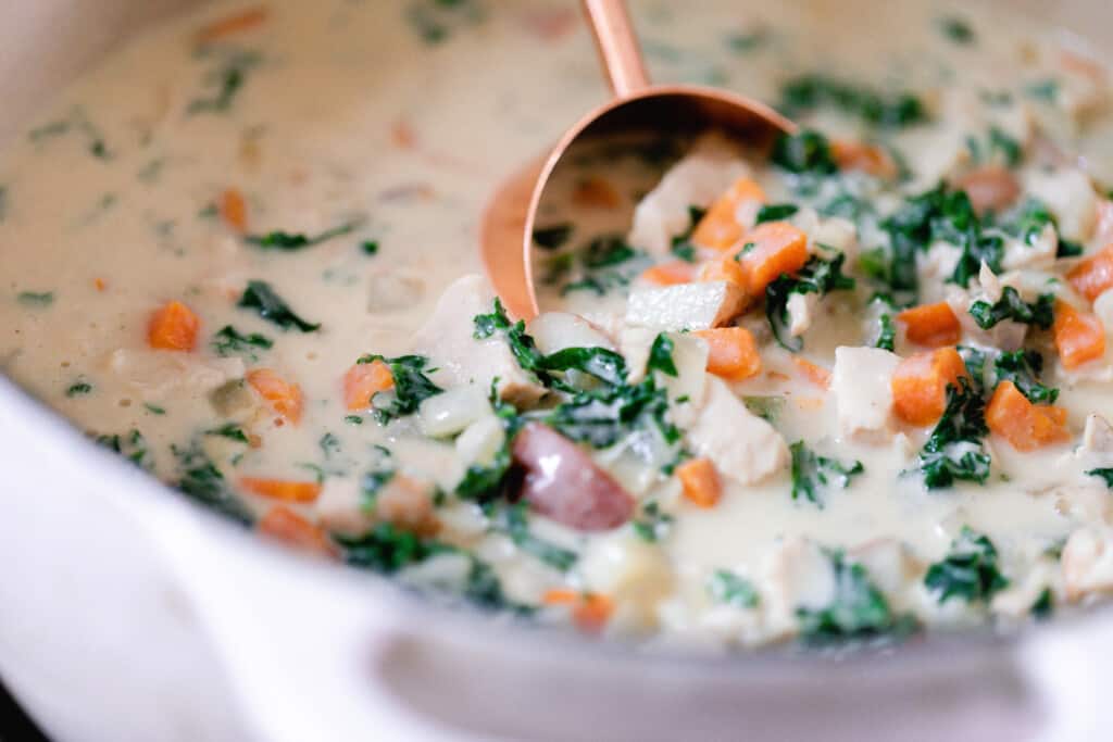 chicken kale soup in a white dutch oven being scooped out with a copper measuring cup