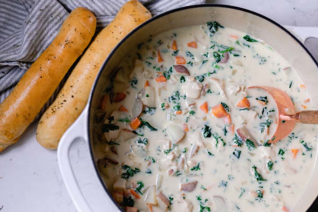 chicken kale soup in a white dutch oven being stirred with wooden spoon with two sourdough breadsticks on the side