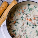 chicken kale soup in a white dutch oven being stirred with wooden spoon with two sourdough breadsticks on the side