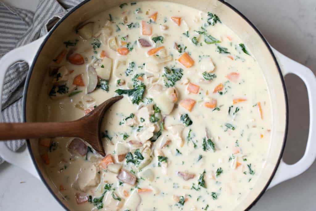 chicken kale soup in a white dutch oven being stirred with a wooden spoon