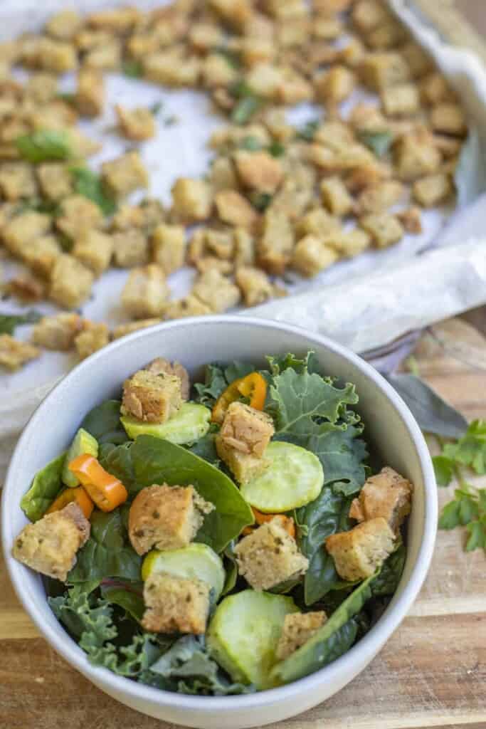a salad topped with homemade gluten free croutons, sliced cucumber and peppers in a bowl with more croutons on a baking pan in the background